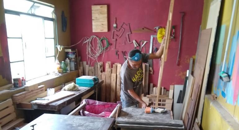 Homem em marcenaria, cercado de materiais como serra, mesa para cortar e outros instrumentos, maneja a madeira de pallets.