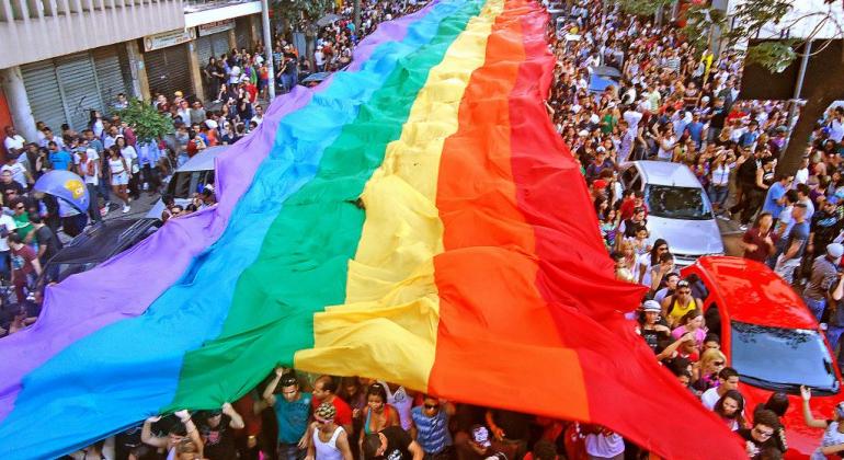 Mais de 800 pessoas em uma rua com uma imensa faixa com as cores do arco-íris, símbolo da luta LGBT, durante o dia.