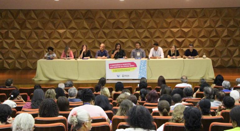 Mesa de abertura da V Conferência Municipal dos Direitos das Pessoas Idosas, com nove integrantes; à frente, mais de 30 pessoas sentadas, assistindo o evento.