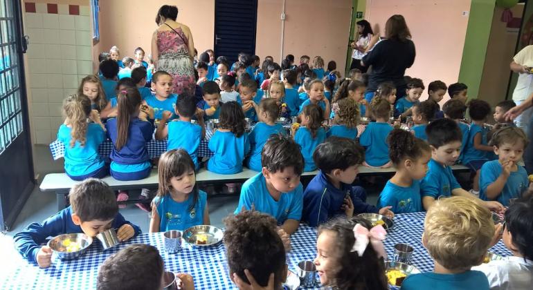 Crianças sentadas em uma mesa para receber alimentação em escola infantil