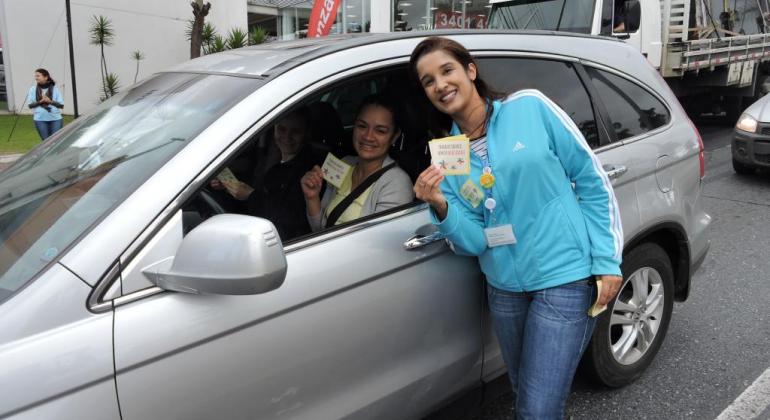 Mulher com panfleto educativo ao lado de carro com motorista, durante o dia. 