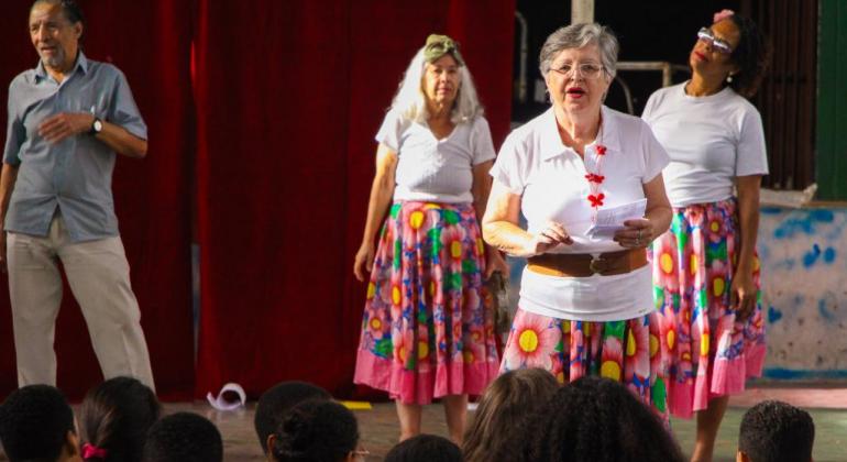 Três senhoras com camisetas brancas e saias de chita se apresentam. 