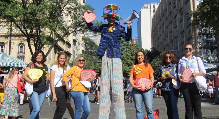 Homem com perda de pau mulheres com a blusa da campanha Maio Laranja posam para foto junto com cidadãos com leques da campanha, no centro da cidade, durante o dia. 