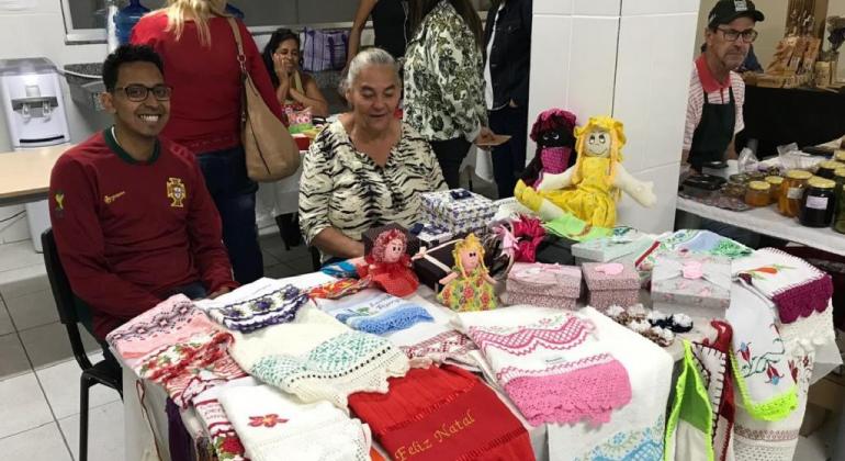 A edição de novembro da Perifeira será realizada nos dias 21 e 22/11, das 16h às 21h, no Centro de Referência da Juventude, no Centro. Foto: Divulgação PBH Jovem e senhora sentados, vendendo panos de prato decorados com crochê sobre a mesa