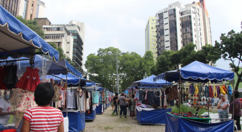 Programa tem como foco principal a formação, promoção, socialização e inclusão produtiva de cidadãos em situação de vulnerabilidade. Foto: Stênio LIma/PBH Mais de seis barraquinhas de lona azul com artesanato e pessoas transitando; ao fundo, dois prédios, durante o dia.