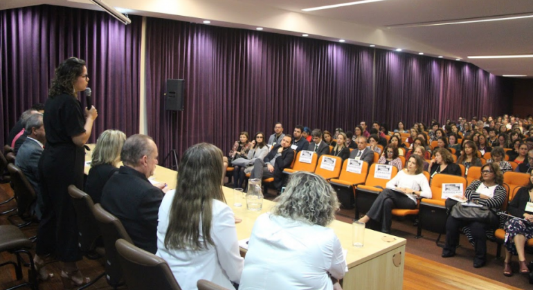 Secretária municipal de Sssistência Social, Segurança Alimentar e Cidadania, Maíra Colares, de pé, ao lado de cinco pessoas sentadas na pesa, falando para mais de trinta pessoas na sala JK, na seda da PBH. 