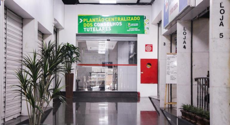 No corredor de um prédio, ao fundo, sala com porta de vidro e os seguintes dizeres: "Plantão Centralizado dos Conselhos Tutelares".