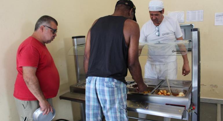 Homem de branco e touca serve comida para dois homens de bermuda. 