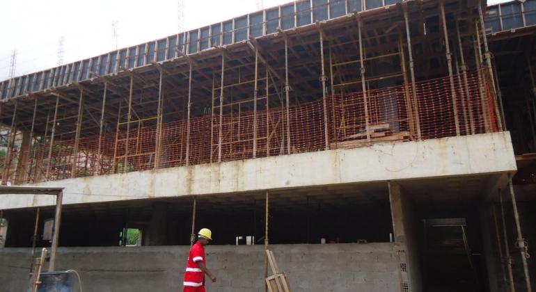 Prédio de dois andares em construção com homem de vermelho e capacete à frente, caminhando. 