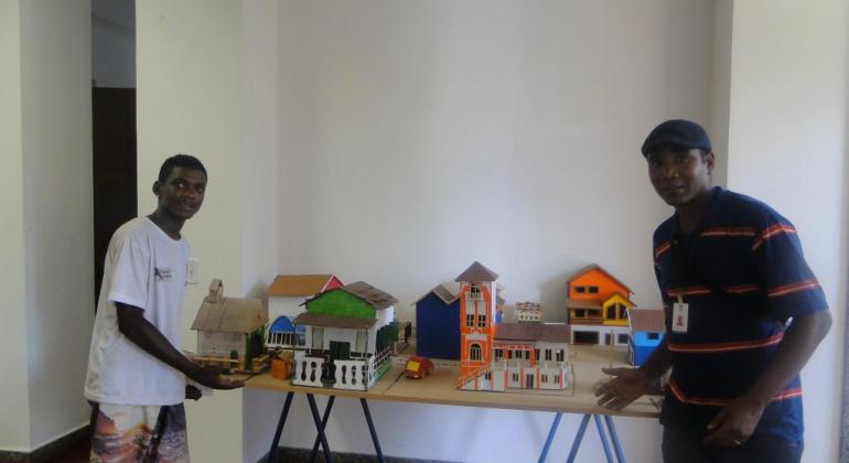 Dois jovens mostram miniaturas de casas construídas, expostas em cima da mesa. 