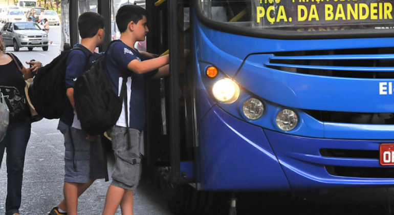 Dois jovens com mochila entram em ônibus; atrás deles está uma mulher. 