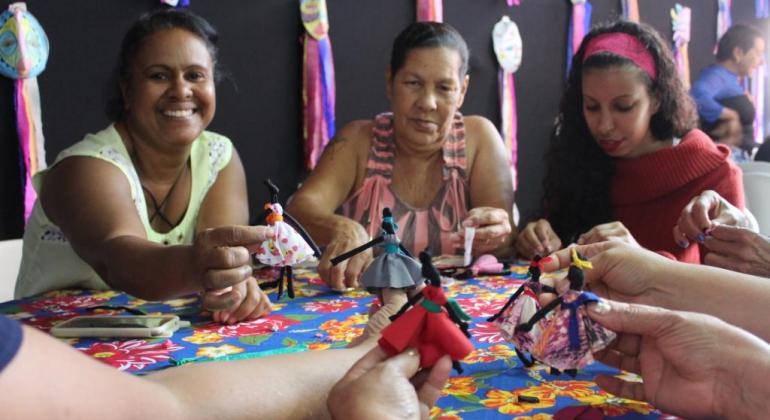 Quatro mulheres mostram as bonequinhas negras - abayomis - em cima da mesa com forro colorido. 