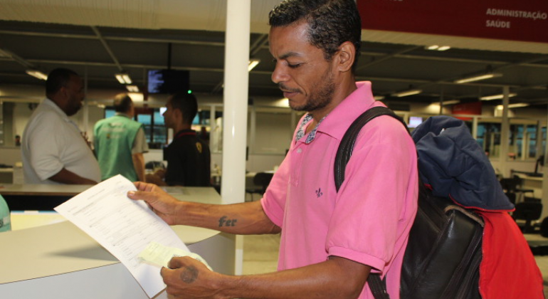 Homem de camiseta pólo rosa e mochila mostra papel, que está em suas mãos. 