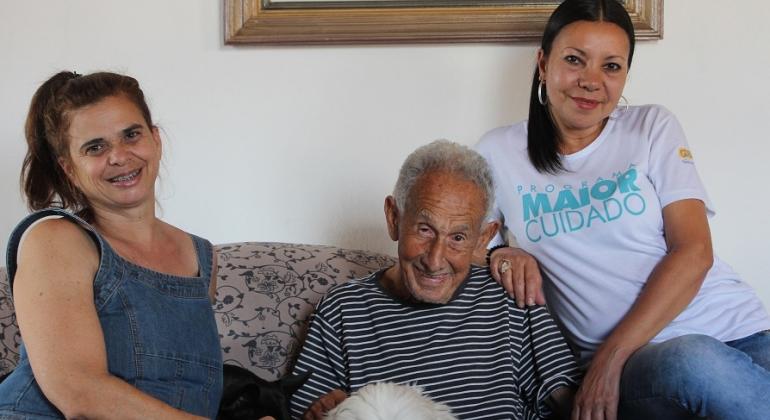 Idoso acompanhado por duas mulheres, uma delas veste uma camiseta com os dizeres? "Maior Cuidado". 