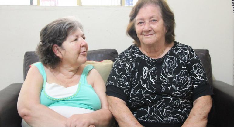 Duas senhoras idosas sentadas em um sofá, em uma sala, durante o dia. 