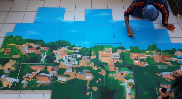 Jovem pinta favela em módulos quadrados no chão. 