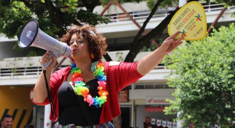 Mulher com megafone e colar colorido de Carnaval mostra leque com os dizeres: "Trabalho infantil nem de brincadeira!"