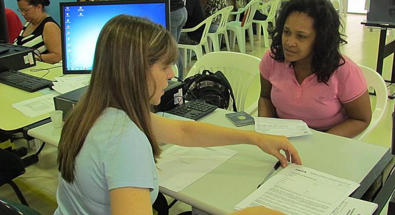 Mulher realiza o atendimento a usuária em uma mesa com outros usuários no plano de fundo