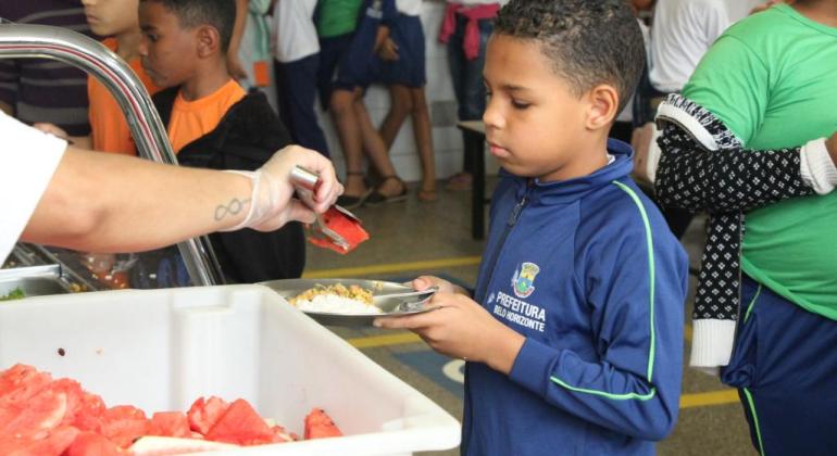 Foto de criança usando uniforme da Prefeitura de Belo Horizonte segurando um prato de comida. Há uma pessoa servindo uma melancia.