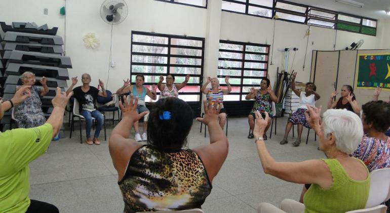Mais de doze adultos, sentados em cadeiras dispostas em forma de círculo e com as mãos para cima, participam de dinâmica pela saúde no Cras Mariano de Abreu. 