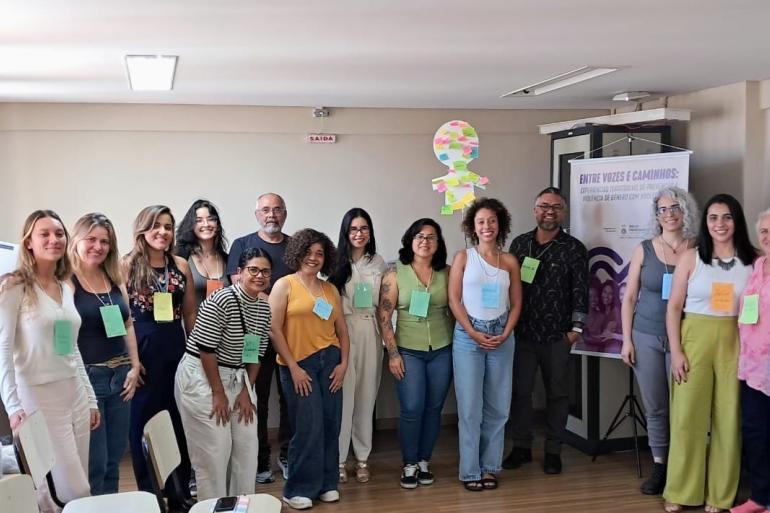 Na foto 13 pessoas estão em pé, próximas e posando para a fotografia. Elas estão em uma sala, com parede bege atrás e teto branco. Ao lado direito, tem um banner.
