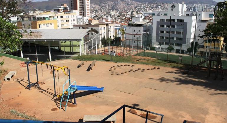 Praça com quadras esportivas coberta e descoberta ao fundo e brinquedos à frente; mais ao fundo, cidade com prédios e montanha, durante o dia. 
