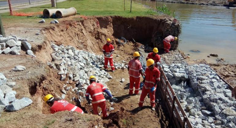 A manutenção das bacias e o controle dos resíduos sólidos realizados pela Sudecap contribuem para evitar o alagamento dos córregos. Foto: Divulgação PBH Seis funcionários da Sudecap, com capacete, fazendo manutenção de resíduos.