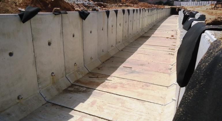 Obras da PBH nos córregos Lareira e Marimbondo vão melhorar escoamento d'água e contribuir com a prevenção de enchentes em Venda Nova. Foto: Divulgação PBH Paredes de cimento ao redor de um córrego, urbanizado para prevenção de enchentes em chuvas, durante o dia.