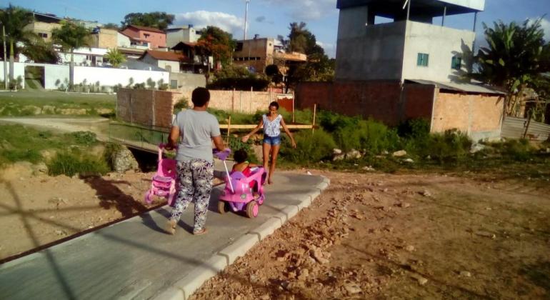 Duas mulheres andam em passeio recém feito; uma delas empurra carrinho rosa com criança e carrega outro carrinho vazio nas mãos. 