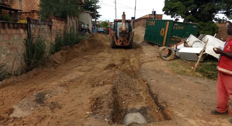 Rua sem asfalto com cano de drenagem sendo colocado no meio e máquina de obras ao fundo, durante o dia. 