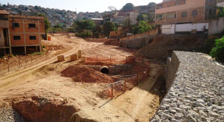 Rua de terra em obras, com brita e canos, durante o dia. 