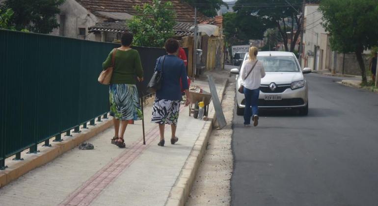 Quatro pessoas andam em calçada ao lado de rua asfaltada, carro transita na rua, durante o dia. 