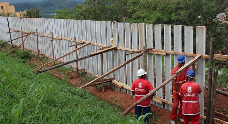 Três funcionários, com capacete e macacão, instalam vigas de concreto que formam uma cerca alta no Abrigo Granja de Freitas, durante o dia. 