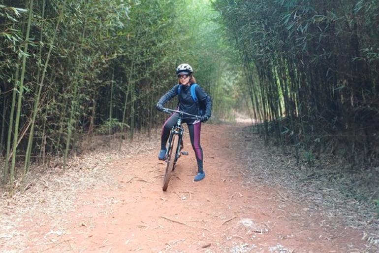 Parque Roberto Burle Marx oferece nova pista de mountain bike no Barreiro Uma mulher vestida com calça legging, blusa de frio, óculos escuro, capacete e carregando uma mochila, em cima de uma bicicleta no meio de uma estrada toda de terra cercada por árvores, ela esta em uma trilha.