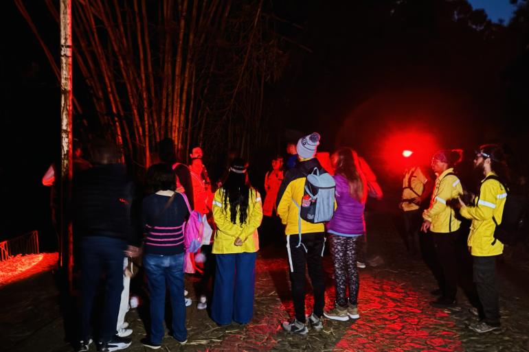Trilha noturna guiada pelo Parque da Serra do Curral terá turma extra na próxima quinta-feira (30) Pessoas reunidas de costas para a câmera, todas vestidas com calças, blusas de frio, tênis esportivo e carregando bolsas e mochilas. A foto foi realizada no período da noite, em local aberto e com muita natureza ao redor. Uma das pessoas carrega uma lanterna de luz vermelha que ilumina toda a região.