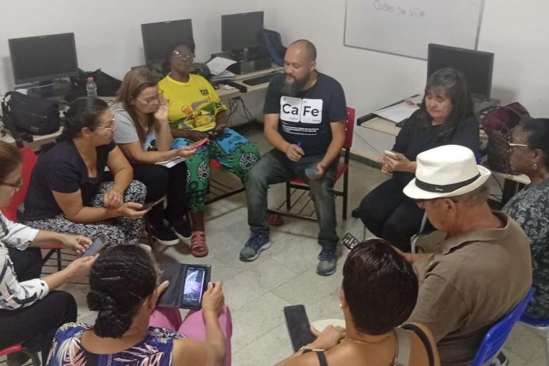 Em uma sala um grupo de pessoas sentam em cadeiras em roda, algumas conversando entre si e outras usando o celular. Atrás das pessoas existem computadores em mesas. 