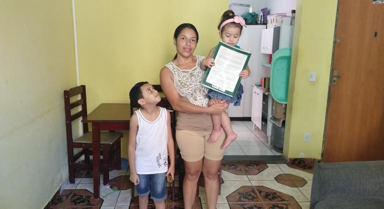 Em dezembro, a Prefeitura de BH, por meio da Urbel, entregou os registros de propriedade em cinco conjuntos regularizados na Capital. Foto: Divulgação PBH Mulher em casa com dois filhos, um deles no colo e com o título de propriedade em mãos.