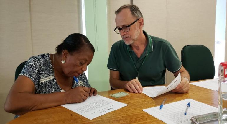 A Urbel está cadastrando as primeiras famílias que serão beneficiadas pelo Programa de Locação Social do Município. Foto: Divulgação PBH Senhora assina papéis, acompanhada por homem de óculos.