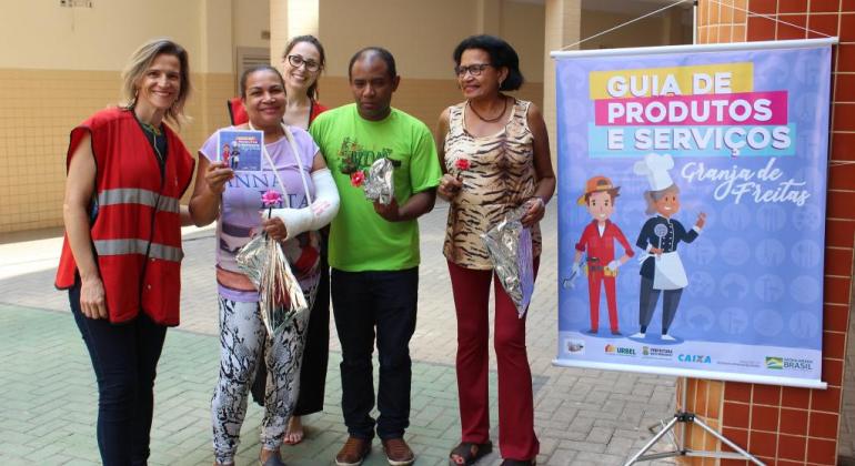 Ao lado de um banner do Guia de Produtos e serviços do Granja de Freitas, duas técnicas da Urbel acompanhada de duas mulheres e um homem, uma delas com o guia na mão. Foto: Divulgação PBH