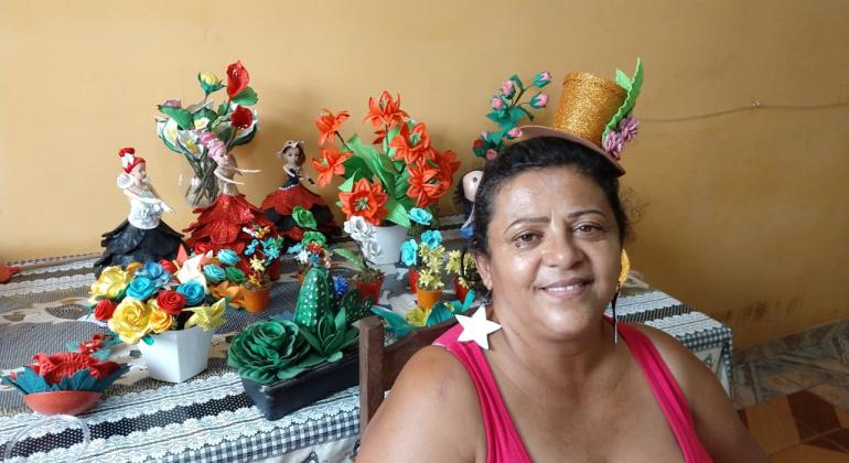 Mulher com chapéu de artesanato, à frente de mesa com arranjos de flores artificiais e bonecas decoradas. 