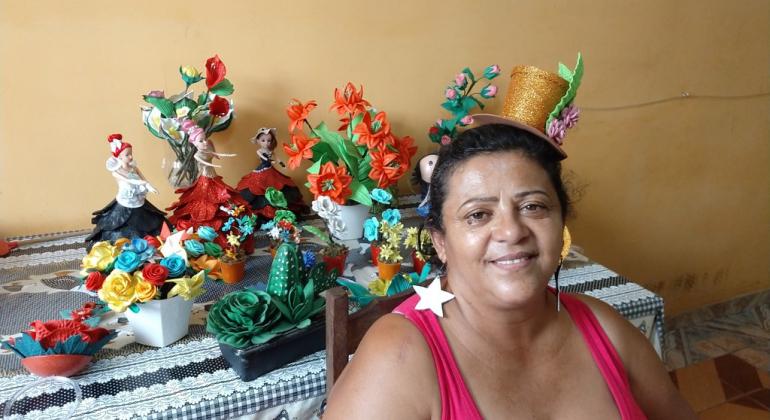 Artesanato, roupas,pintura, e fotografia são alguns dos itens que farão parte do Guia de Produtos e Serviços do Conjunto Granja de Freitas. Foto: Divulgação PBH Mulher com chapéu de artesanato, à frente de mesa com arranjos de flores artificiais e bonecas decoradas.