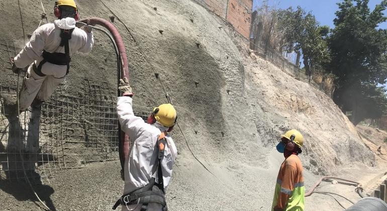 A Urbel retomou, em 2019, uma obra de contenção da encosta que existia no local, beneficiando cerca de 40 famílias das proximidades. Foto: Divulgação PBH Em frente a uma encosta cimentada, três homens trabalham, com capacetes de proteção, durante o dia.