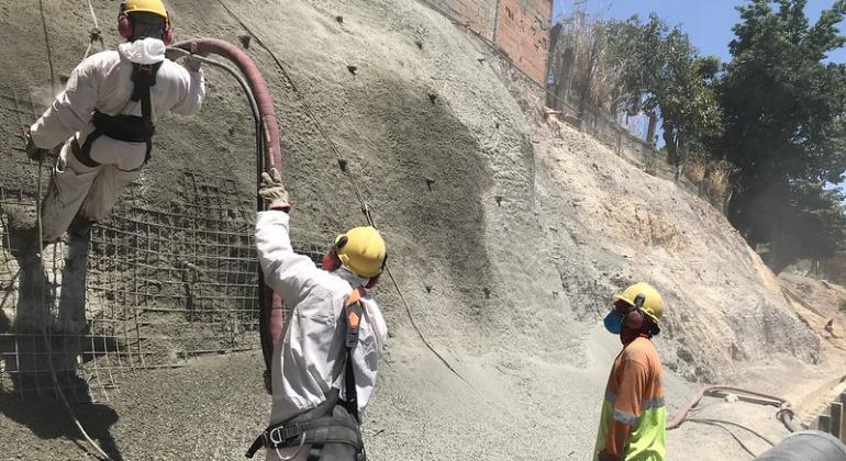 Em frente a uma encosta cimentada, três homens trabalham, com capacetes de proteção, durante o dia.