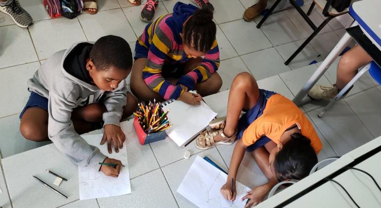 Três crianças sentadas no chão fazem desenhos em papel branco com lápis de cor