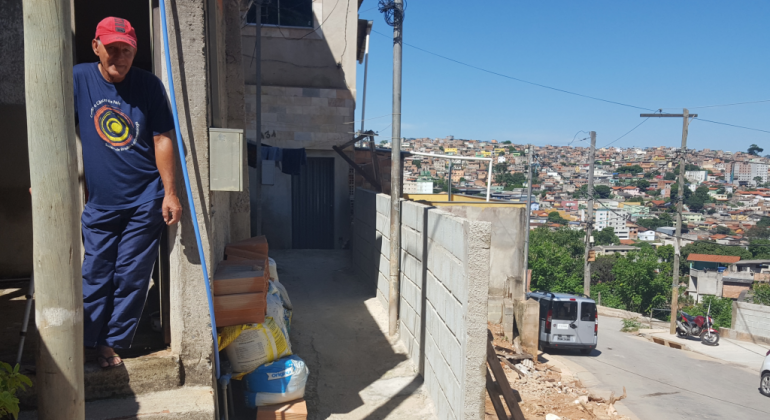 Homem de boné vermelho e camiseta azul, ao lado de rua com obras de urbanização, durante o dia. 