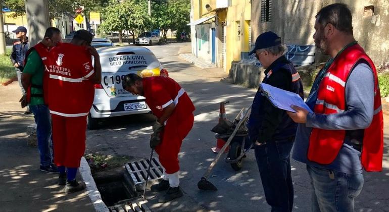 Cinco funcionários e técnicos conferem se bueiro está desentupido em rua de Belo Horizonte, durante o dia. 