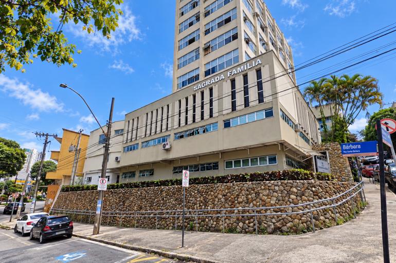 Prefeitura abre licitação para obras de drenagem e reforma da URS Sagrada Família Foto horizontal da fachada do edifício Sagrada Família, em Belo Horizonte, com vários andares, janelas, árvores, muro de pedras, calçada, carros e sinalização de trânsito.