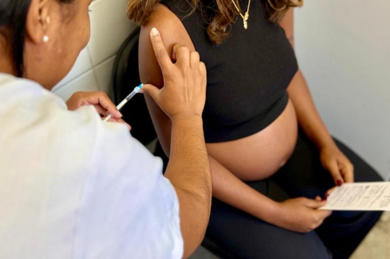 A foto mostra uma gestante recebendo uma vacina no braço aplicada por uma profissional de saúde. 