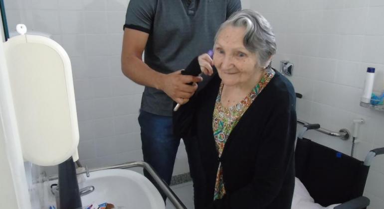 Paciente idosa treina movimentos cotidianos em frente a uma pia, auxiliada por técnico do Centro de Reabilitação de Venda Nova.