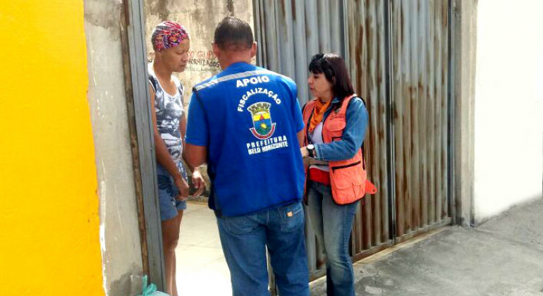 Dois técnicos da prefeitura abordam mulher no portão da sua casa, durante o dia. 